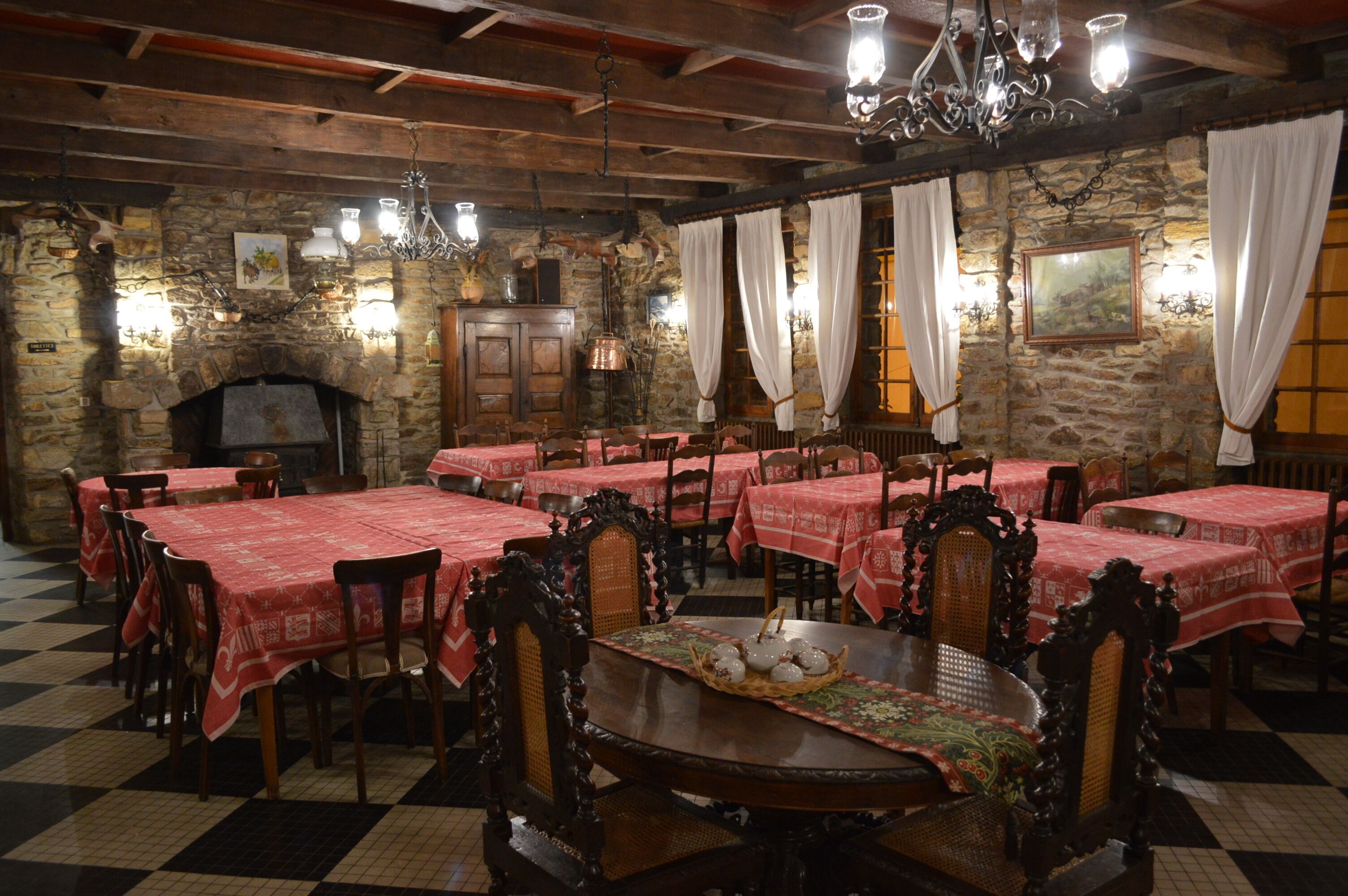 A restaurant with stone walls, wooden beams, and red tablecloths. Tables are arranged around a central fireplace.