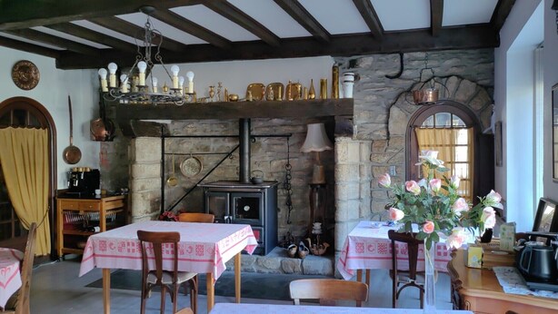 Une salle à manger rustique avec une cheminée en pierre, des poutres en bois et un lustre. Les tables sont dressées avec des nappes roses et des chaises.