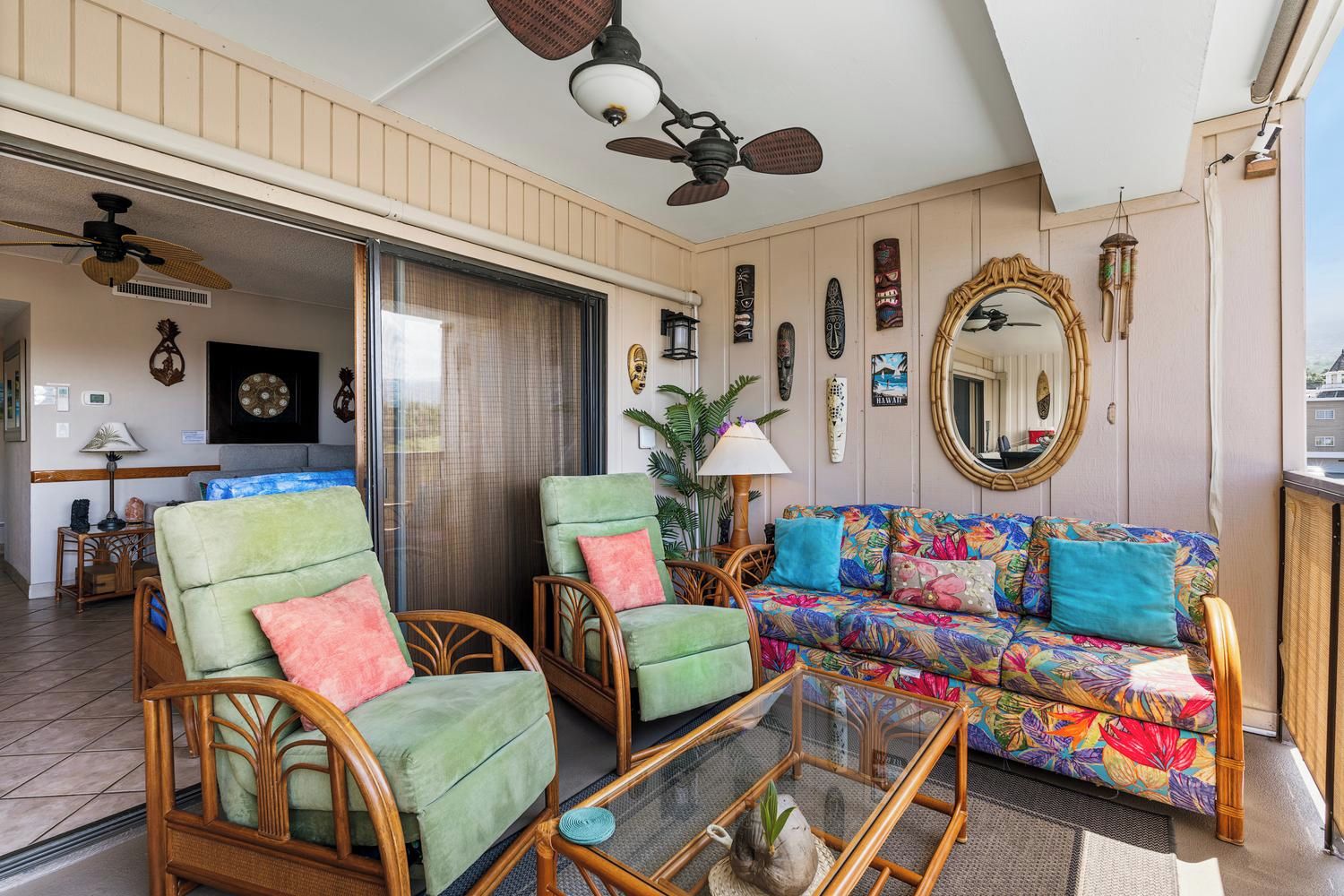 A room with wicker chairs, a colorful sofa, a glass coffee table, and a ceiling fan.