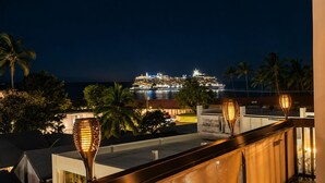 Vista noturna de um navio de cruzeiro atracado em um porto, com palmeiras e um edifício ao primeiro plano.