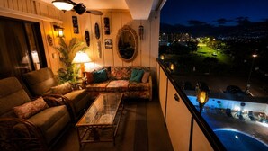 Uma varanda com sofá, cadeiras, mesa e vista para uma piscina e paisagem urbana à noite.