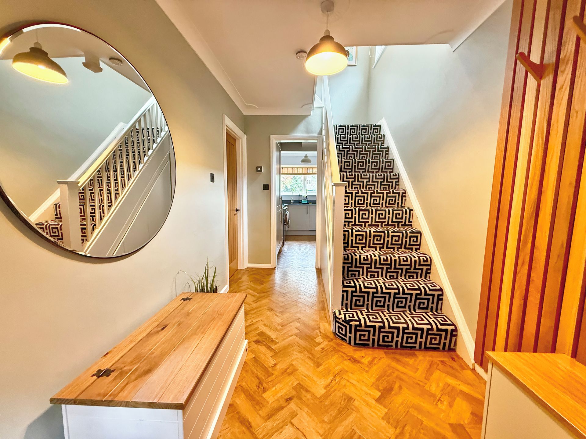 A hallway with a wooden bench, a mirror, a staircase with patterned steps, and a view into a kitchen area.