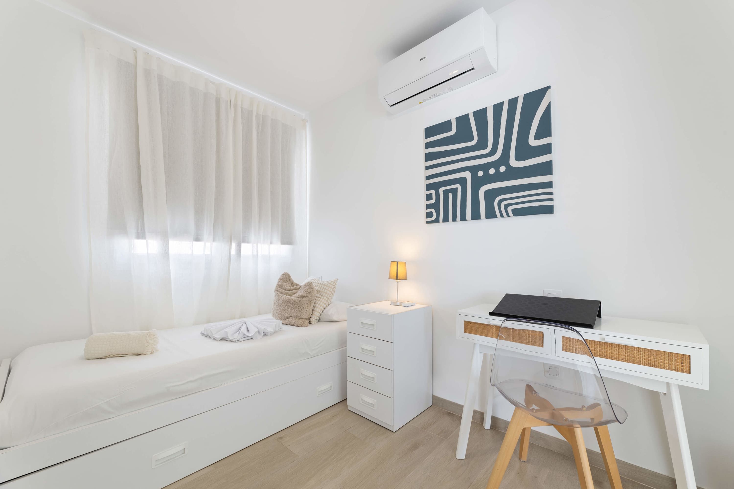 Un dormitorio minimalista con cama, mesita de noche, lámpara y un escritorio con silla.