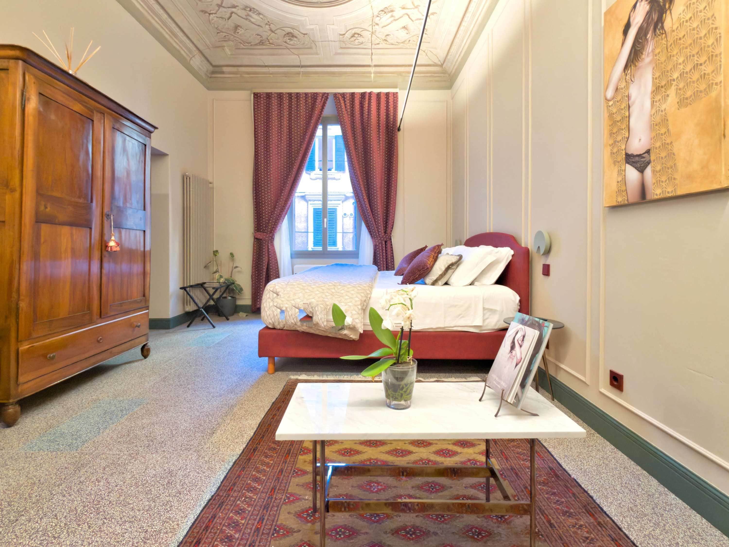 Une chambre avec une grande armoire en bois, un lit avec une literie rouge, une table blanche ornée d’un vase de fleurs et un tapis à motifs.