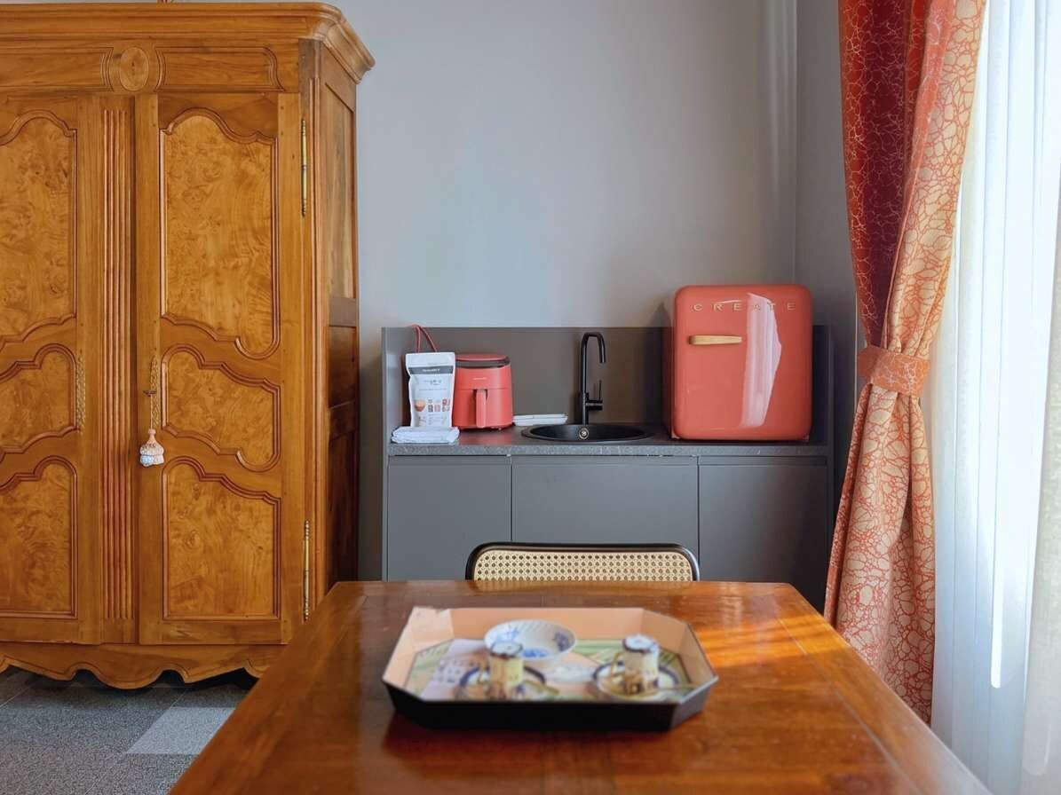 Un meuble en bois, une petite cuisine équipée avec un appareil électroménager rouge, et une table sur laquelle se trouve un plateau rempli d’objets.