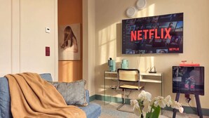 Un salón con un sofá azul, una mesita de centro, un televisor mostrando el logo de Netflix, un escritorio pequeño con una lámpara y una estantería de pared.