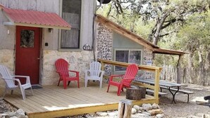 A rustic stone building with a red door, a wooden deck, and several Adirondack chairs. - Hideaway Cabin 3 Bedroom Cabin (Uvalde)