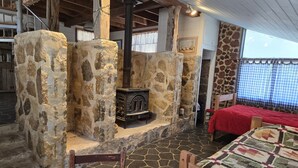 A rustic room with a stone fireplace, wooden beams, a bed with a red bedspread, and a quilted tablecloth. - Hideaway Cabin 3 Bedroom Cabin (Uvalde)