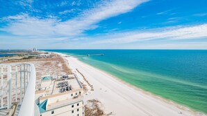 A beach with a pier extending into the ocean, a clear sky, and a building with a green roof in the foreground.