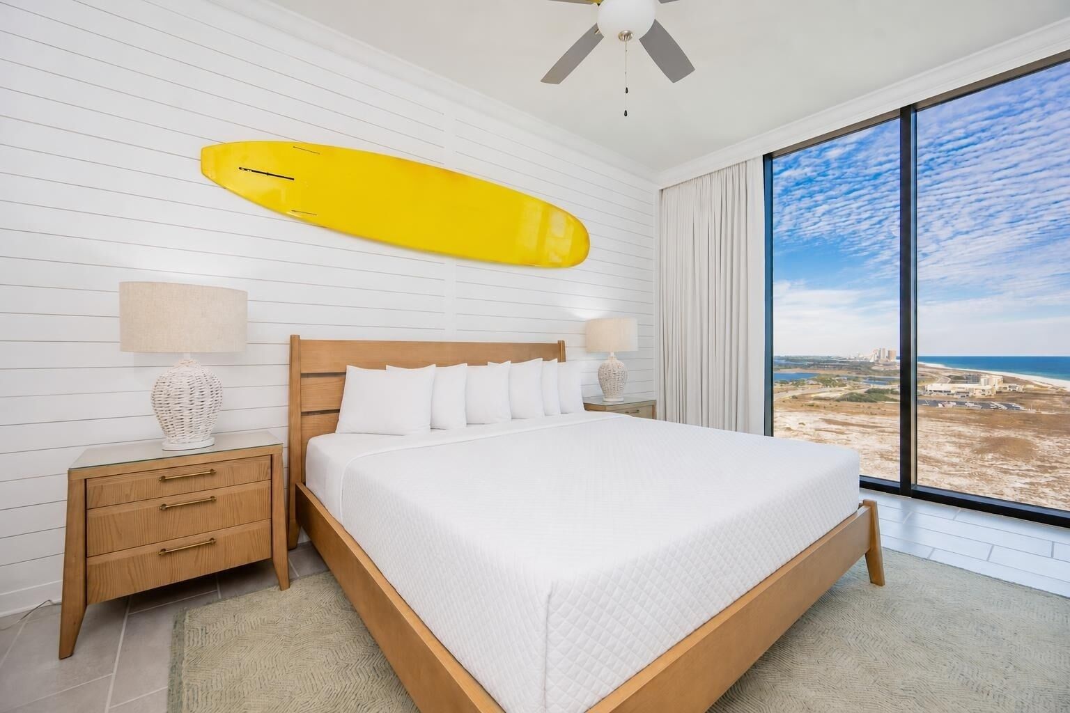 A bedroom with a large bed, wooden headboard, two bedside tables with lamps, a ceiling fan, and a large window with a beach view.