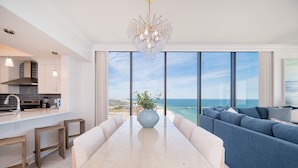 A modern kitchen with a large island, a dining area with a glass table, and a living area with a sofa and a view of the ocean.