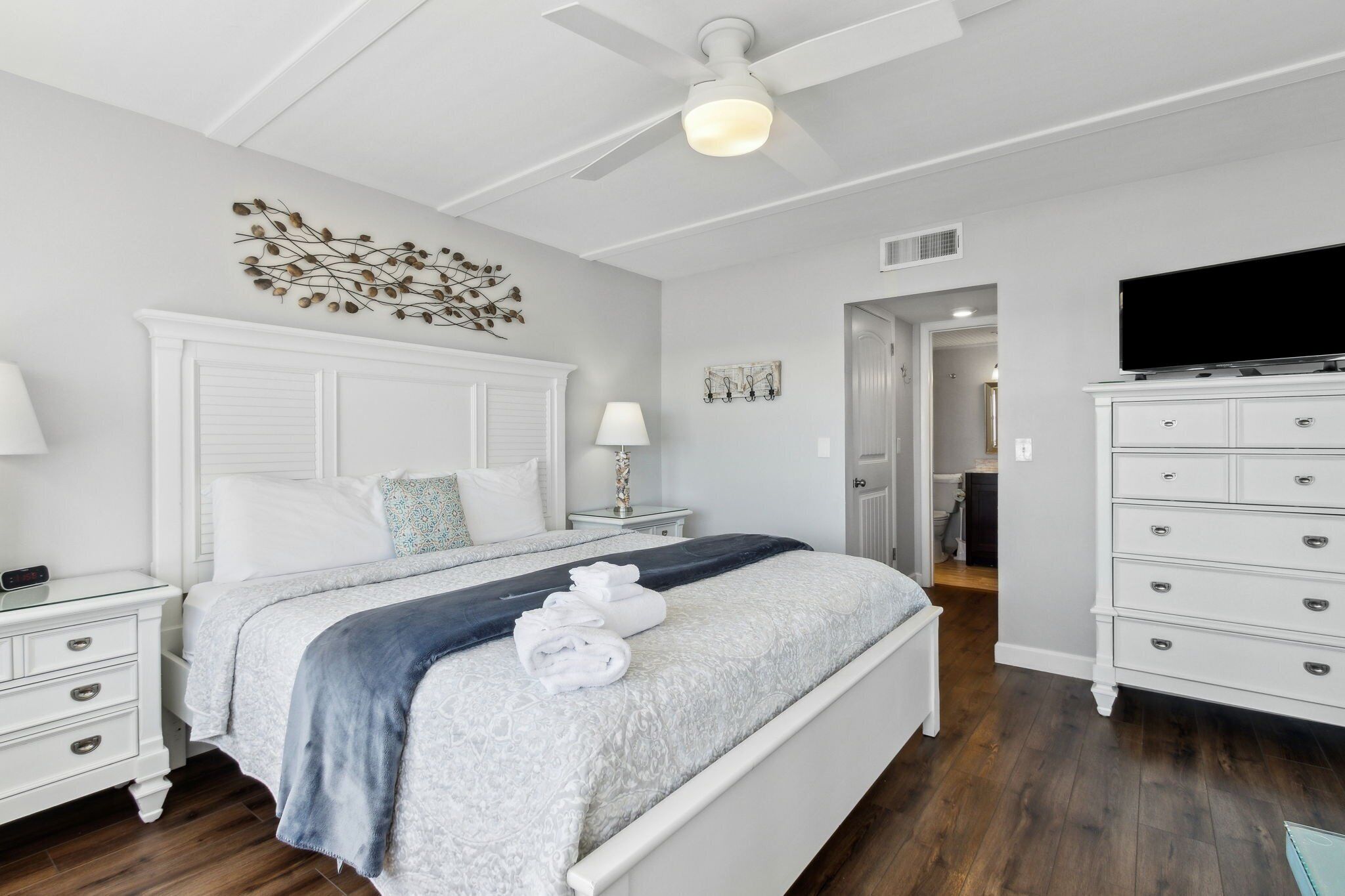 Une chambre à coucher comprenant un lit, deux tables de chevet blanches, une commode, un ventilateur de plafond et un téléviseur.