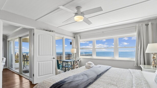 A bedroom with a bed, a ceiling fan, a window with a view of the sea, and a balcony with a table and chairs.