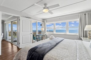Een slaapkamer met een bed, een plafondventilator, een raam met uitzicht op zee en een balkon met een tafel en stoelen.