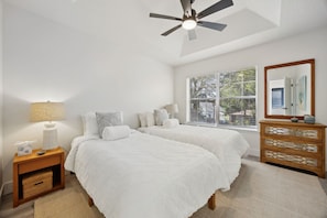 Quarto com duas camas, mesa de cabeceira, c ômoda, ventilador de teto e janela com persianas.
