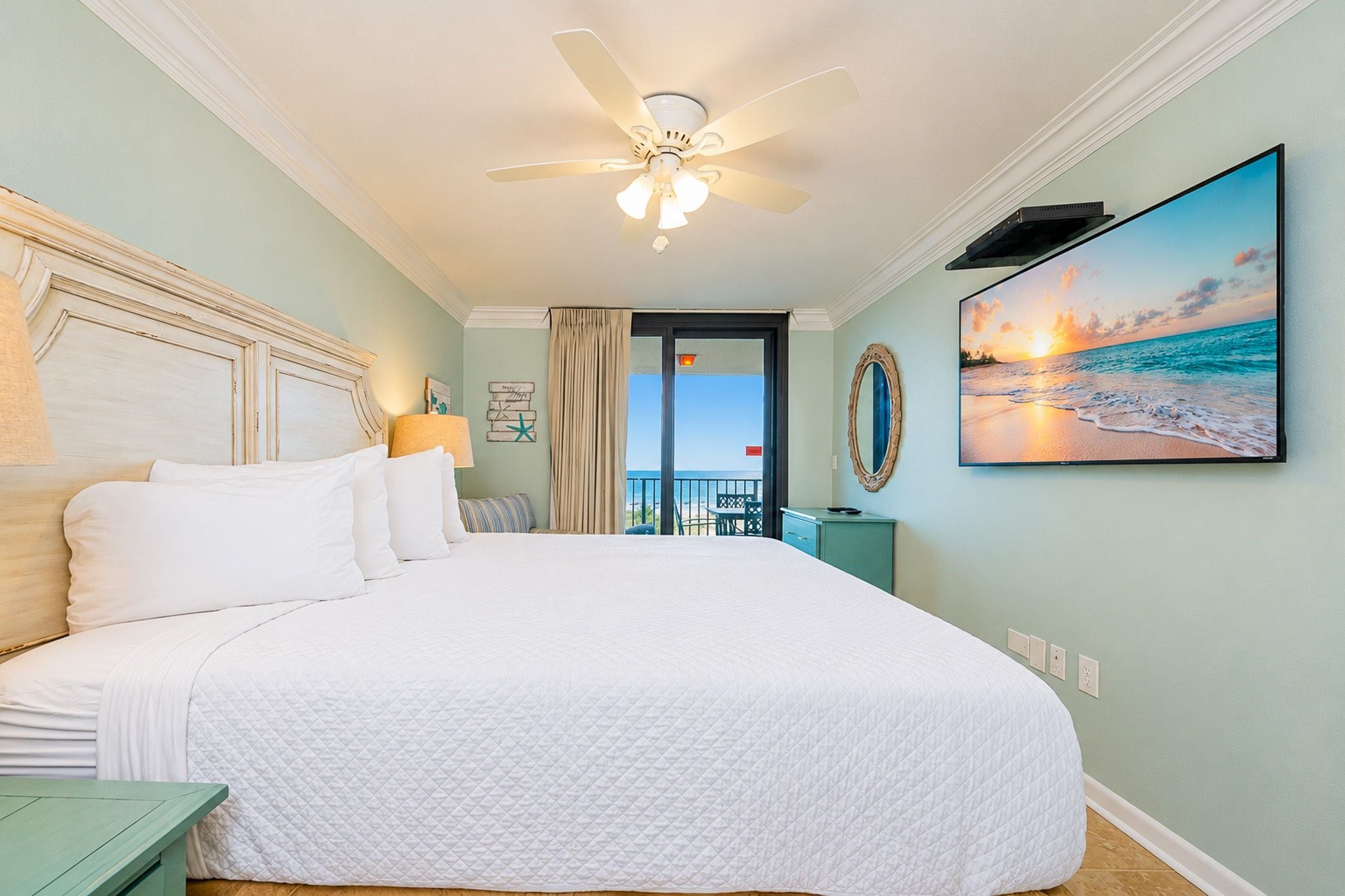 A spacious bedroom with a large bed, a ceiling fan, a view of the ocean through a balcony, and a painting of a sunset.