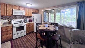 Une cuisine compacte avec des meubles en bois, un four à micro-ondes, une cuisinière et un réfrigérateur. On y trouve une table à manger avec des chaises et une fenêtre avec des stores.