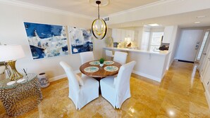 A dining area with a round table, white chairs, and a chandelier. There are abstract paintings on the wall and a kitchen in the background.