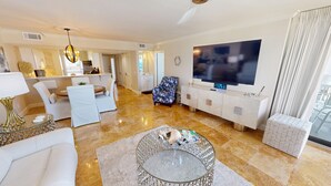 A modern living room with a large TV, a dining area with a white table and chairs, and a kitchen in the background.