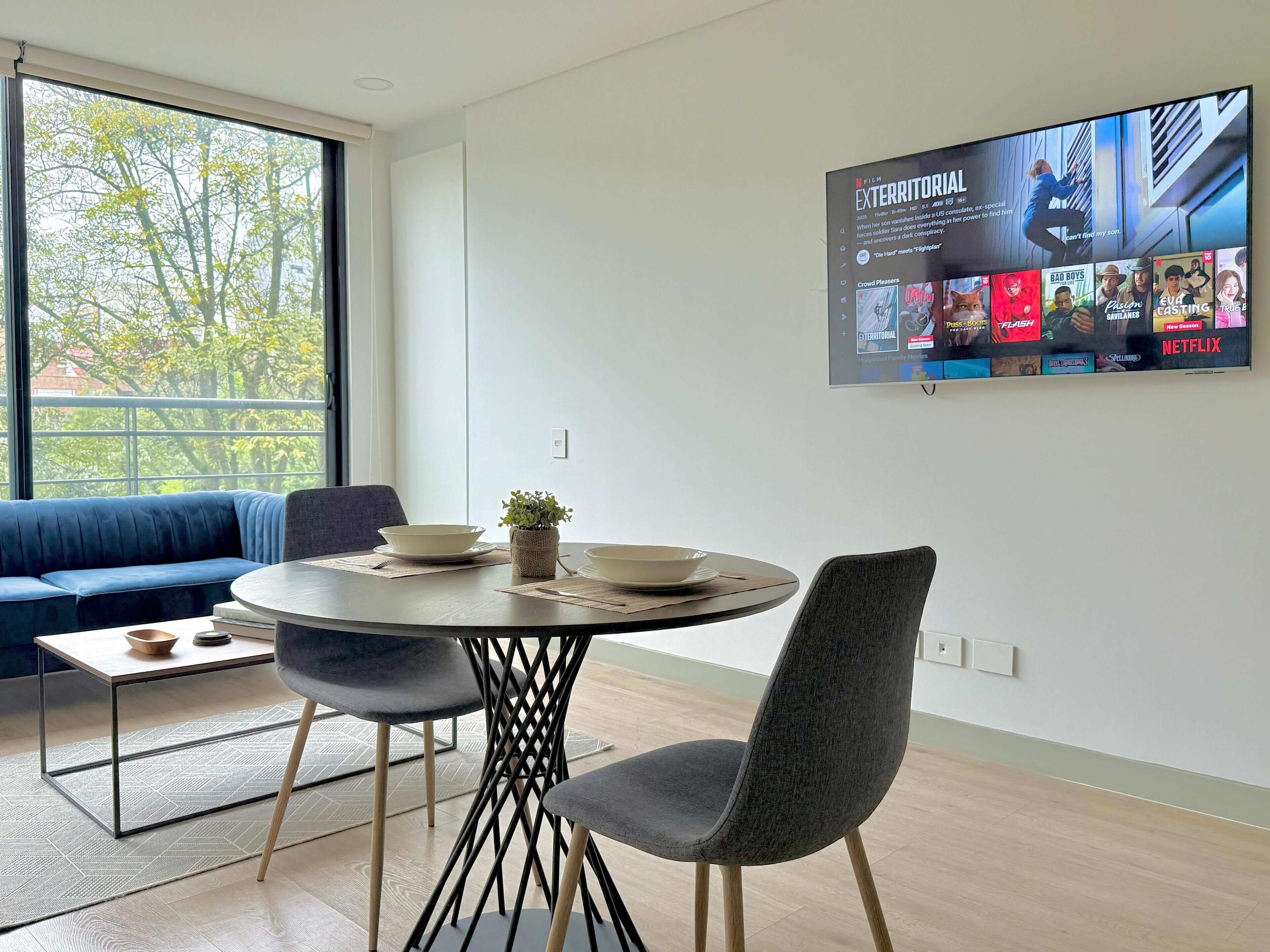 A modern living room with a round dining table, two chairs, a blue sofa, and a TV mounted on the wall displaying a film.