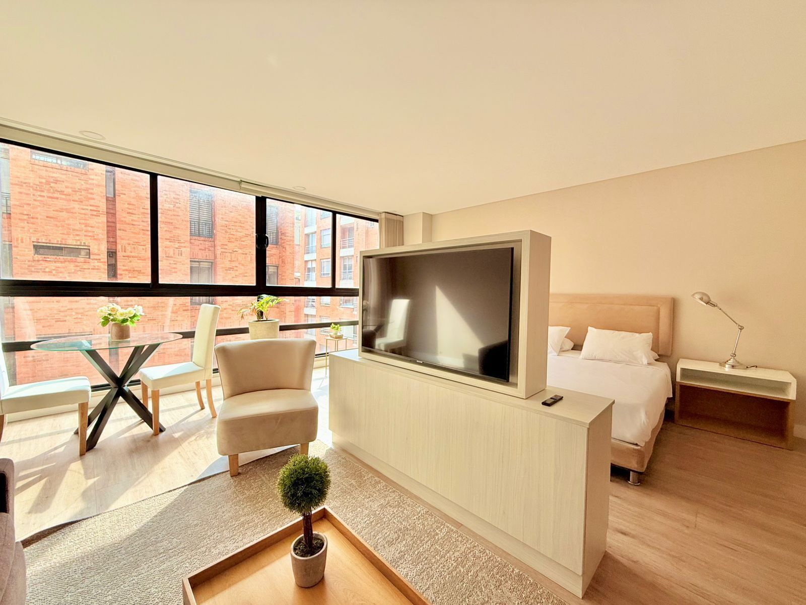 A modern hotel room with a large TV, a bed, a desk with a chair, a small potted plant, and a view of a brick building through large windows.