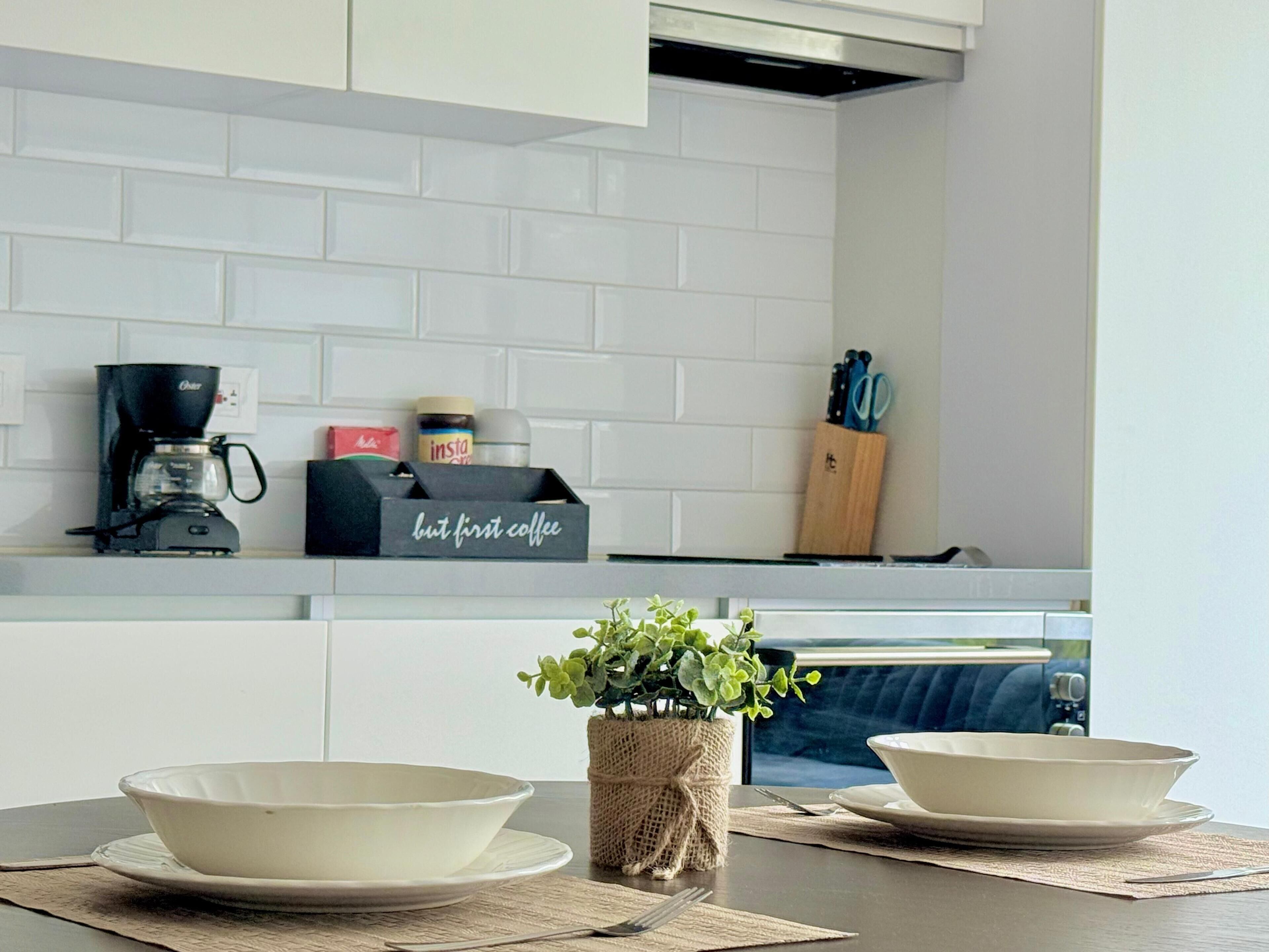 Een moderne keuken met witte kastjes, een koffiezetapparaat, een doosje met de tekst 'eerst koffie' en een plant op het aanrecht.