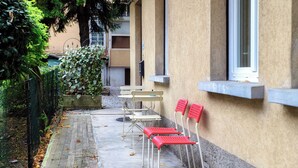 An outdoor seating area with tables and chairs, a concrete pathway, and a building with a window and a door. - Como Gateway (Como)