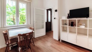 A room with a white dining table and chairs, a white TV stand with a flat-screen TV, and a view of greenery through a window. - Como Gateway (Como)