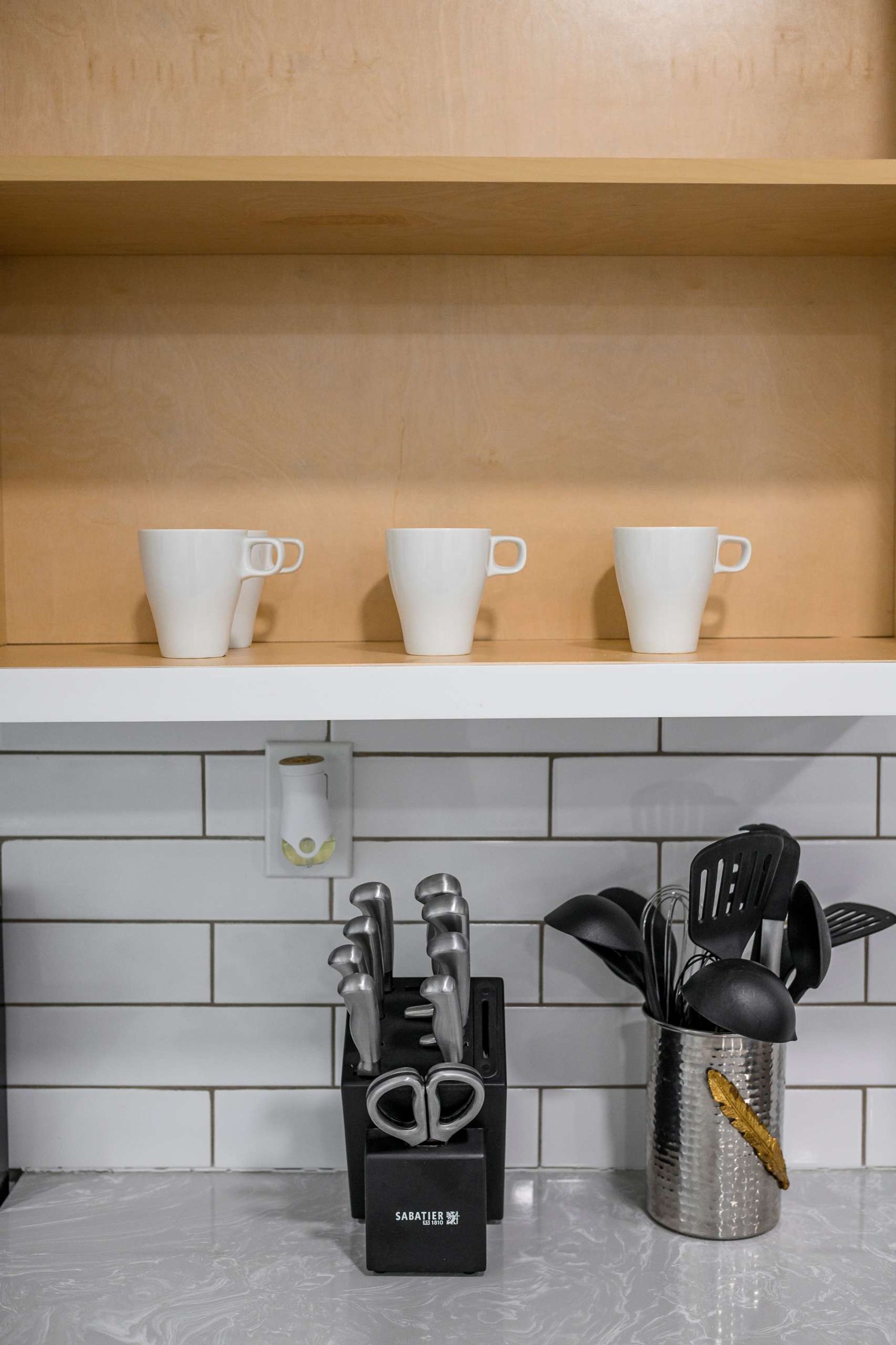 A kitchen shelf with three white cups and a knife block with utensils.