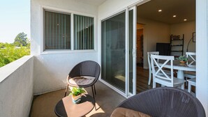 A balcony with a chair, a small table with a plant, and a sliding glass door leading to a dining area with white chairs and a table. - Stylish Home w Free Parking Near LA Hotspots 8 (Altadena)
