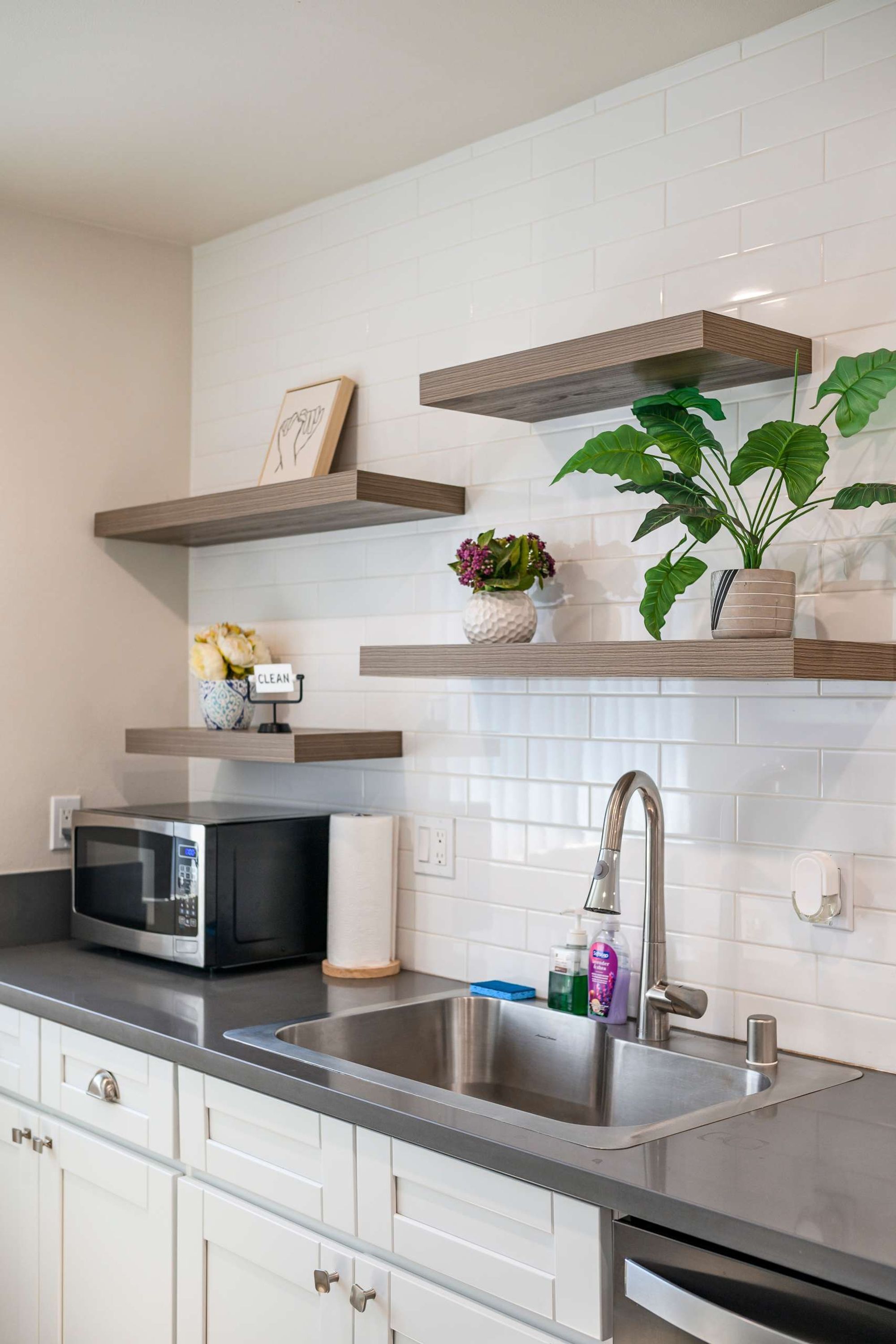 Une cuisine moderne avec des meubles blancs, un évier en acier inoxydable, un four à micro-ondes et des étagères en bois ornées de plantes et d’objets décoratifs.