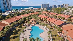 An aerial view of a resort with a central swimming pool, surrounded by resort buildings with red roofs, palm trees, and a view of the ocean and city skyline in the background. - Gulf Highlands escape - pool and beach access (Panama City Beach)