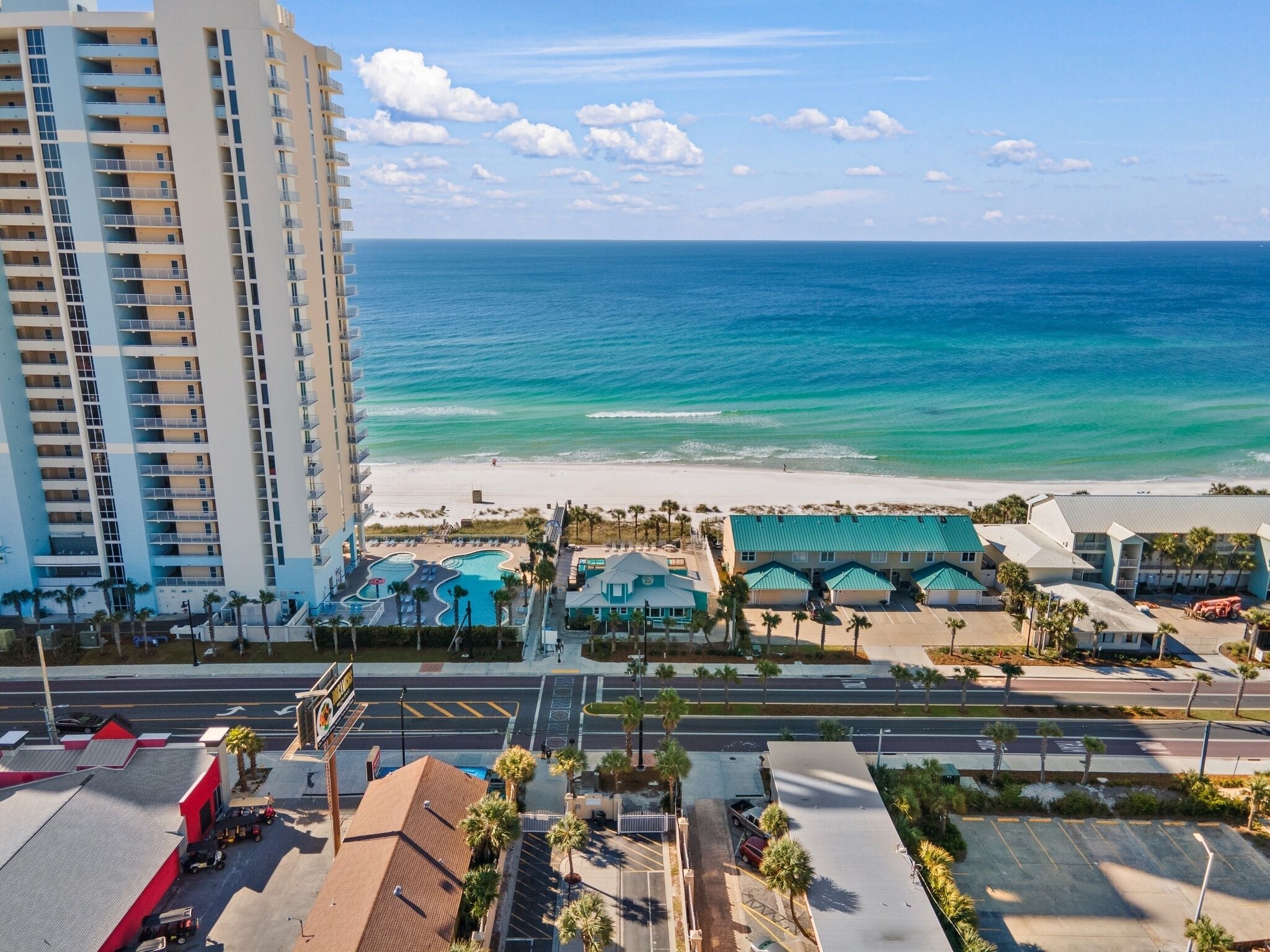 A high-rise hotel overlooking a beach with a pool area and a parking lot.