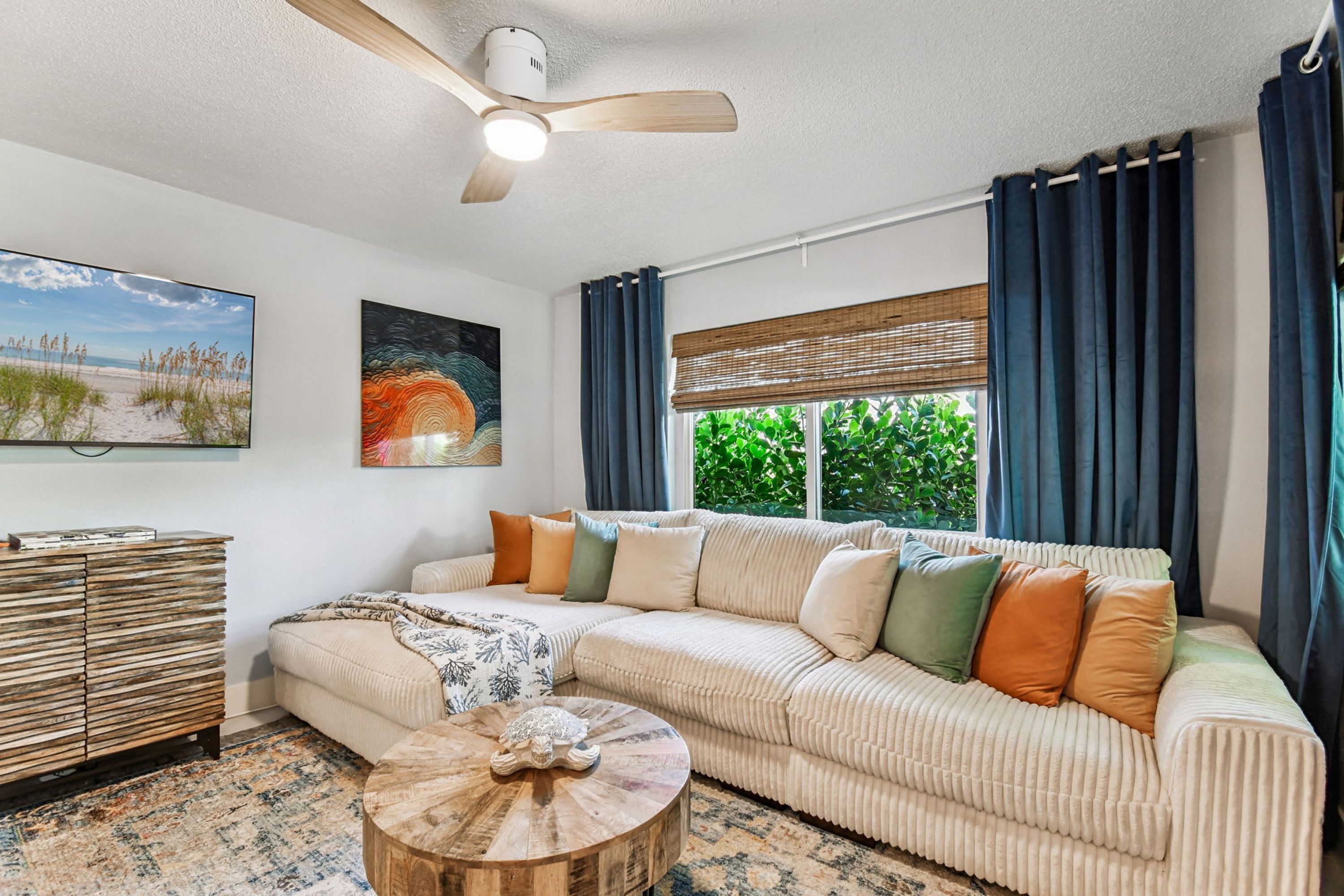 Ein Wohnzimmer mit einem großen, beigefarbenen Ecksofa, einem Holztisch, einem Deckenventilator und einem großen Fenster mit Vorhängen.