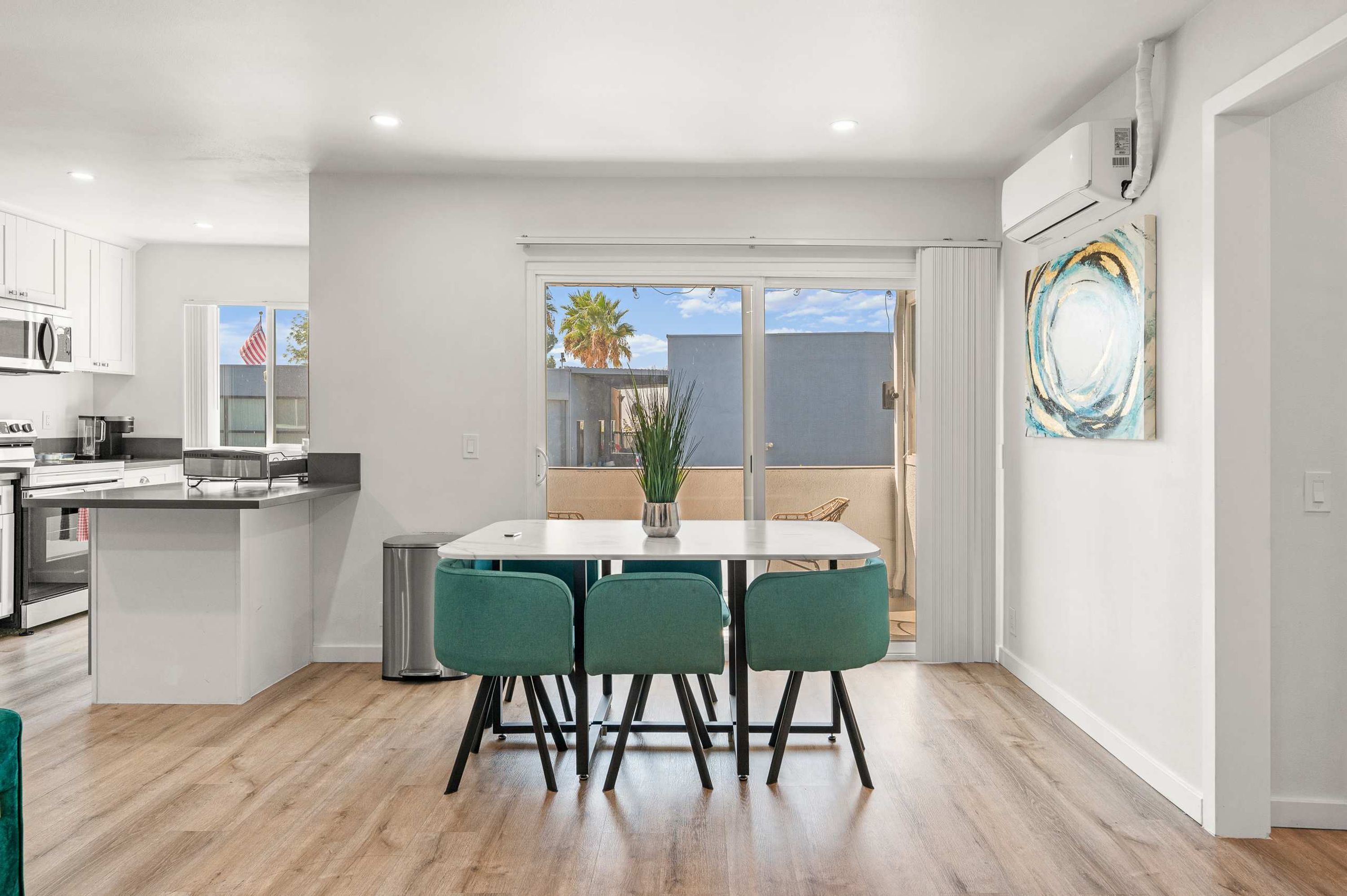 A modern kitchen with white cabinets, a dining area with a white table and green chairs, and a large sliding glass door leading to a balcony with a view of palm trees.