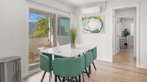 Un intérieur moderne avec un coin repas comprenant une table blanche et des chaises vertes, une grande peinture abstraite au mur et une vue sur un balcon extérieur, accessible par une porte-fenêtre coulissante.