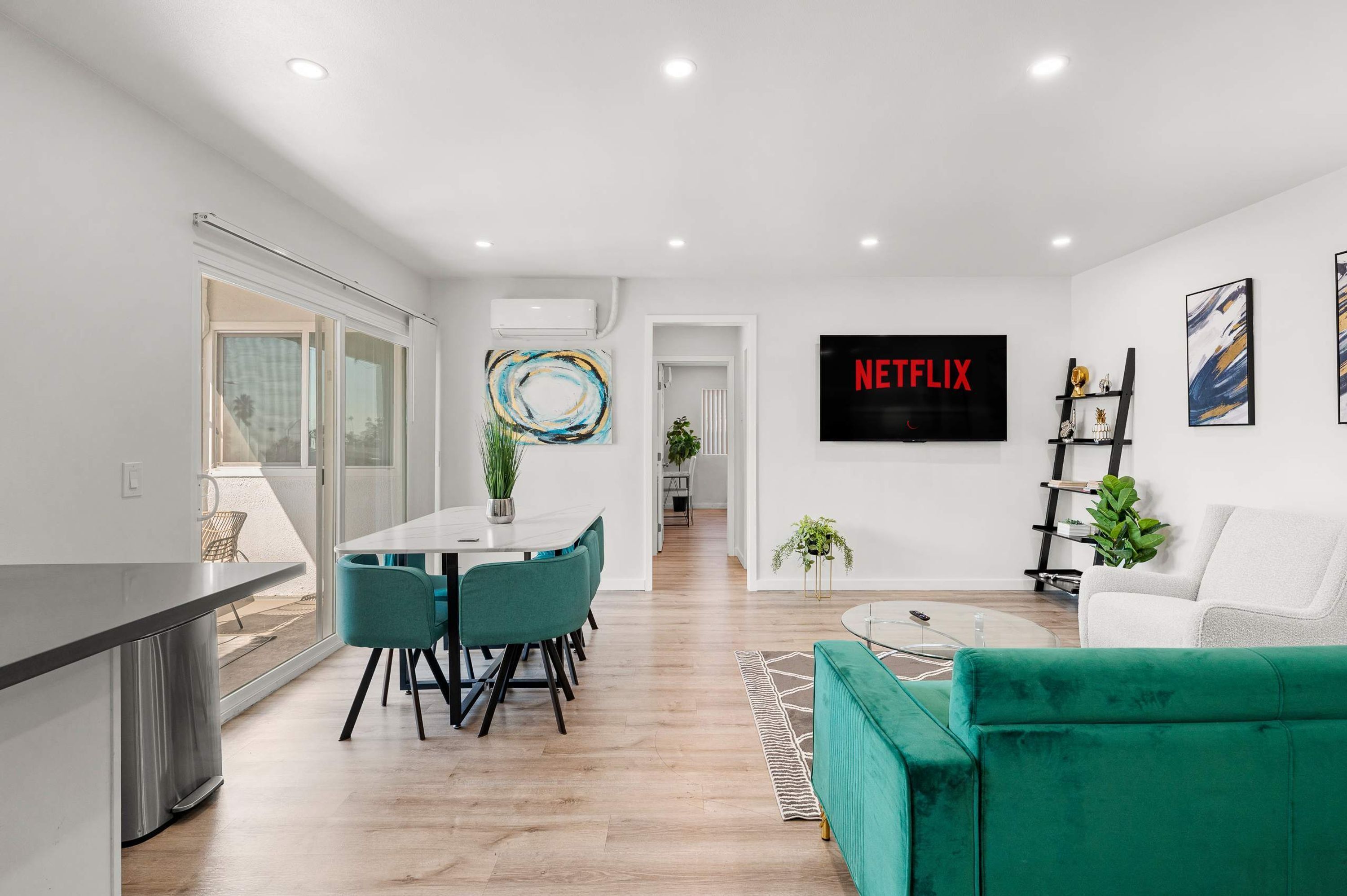 A modern living room with a green sofa, a dining area with teal chairs, a flat-screen TV, and artwork on the walls.