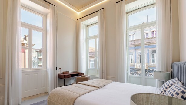 Ein Hotelzimmer mit einem großen Bett, einem Nachttisch, einer Lampe und Blick auf die Stadt durch die Fenster.
