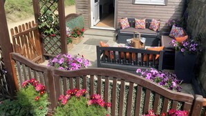 A patio area with a wooden bench, a sofa with colorful cushions, and a wooden pergola. - 3 bedroom cottage in Sark, on the Avenue. (Guernsey)