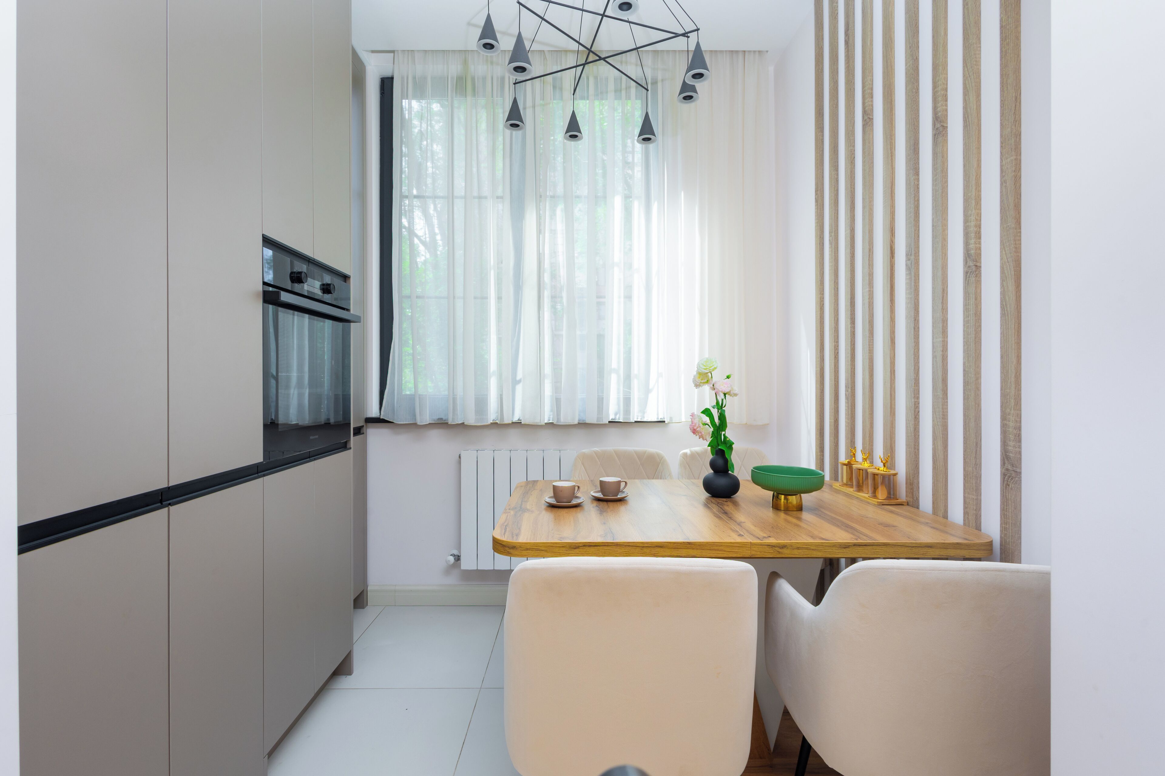 A compact kitchen with a built-in oven, a dining table with chairs, and a window with sheer curtains.