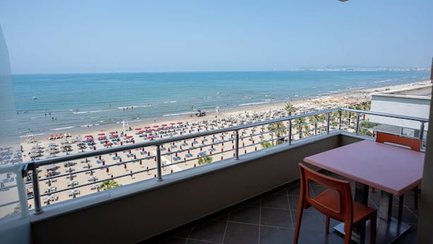 A balcony with a view of a crowded beach, a table, and chairs. - Rumar Hotel (Durres)