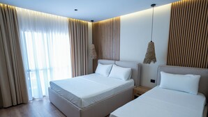 A hotel room with two beds, a wooden floor, a large window with curtains, and a hanging light fixture. - Rumar Hotel (Durres)
