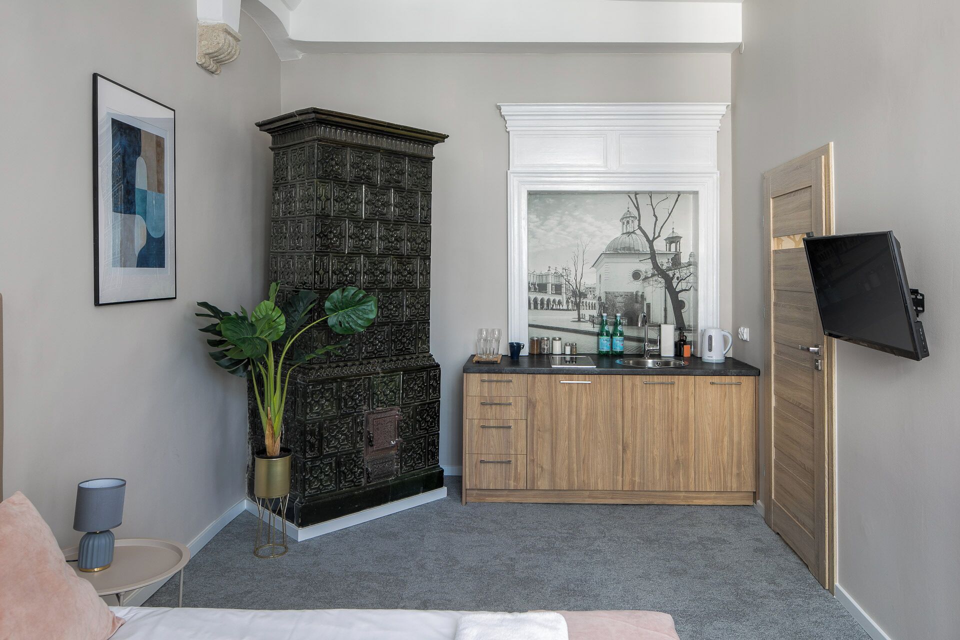 A compact room with a kitchenette, a large window with a cityscape view, a potted plant, and a flat-screen TV mounted on the wall.