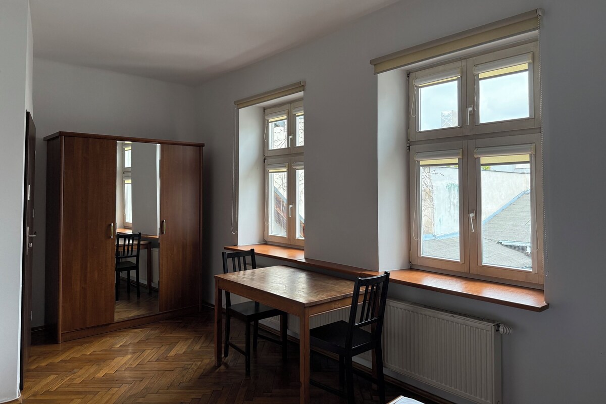 A room with a wooden table and chairs, a wardrobe, and large windows with white frames.
