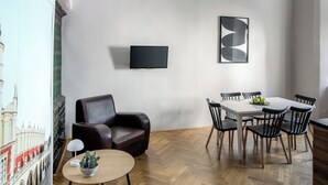 A modern living room with a leather armchair, a round coffee table with a cactus, a dining area with a white table and black chairs, and a flat-screen TV mounted on the wall.