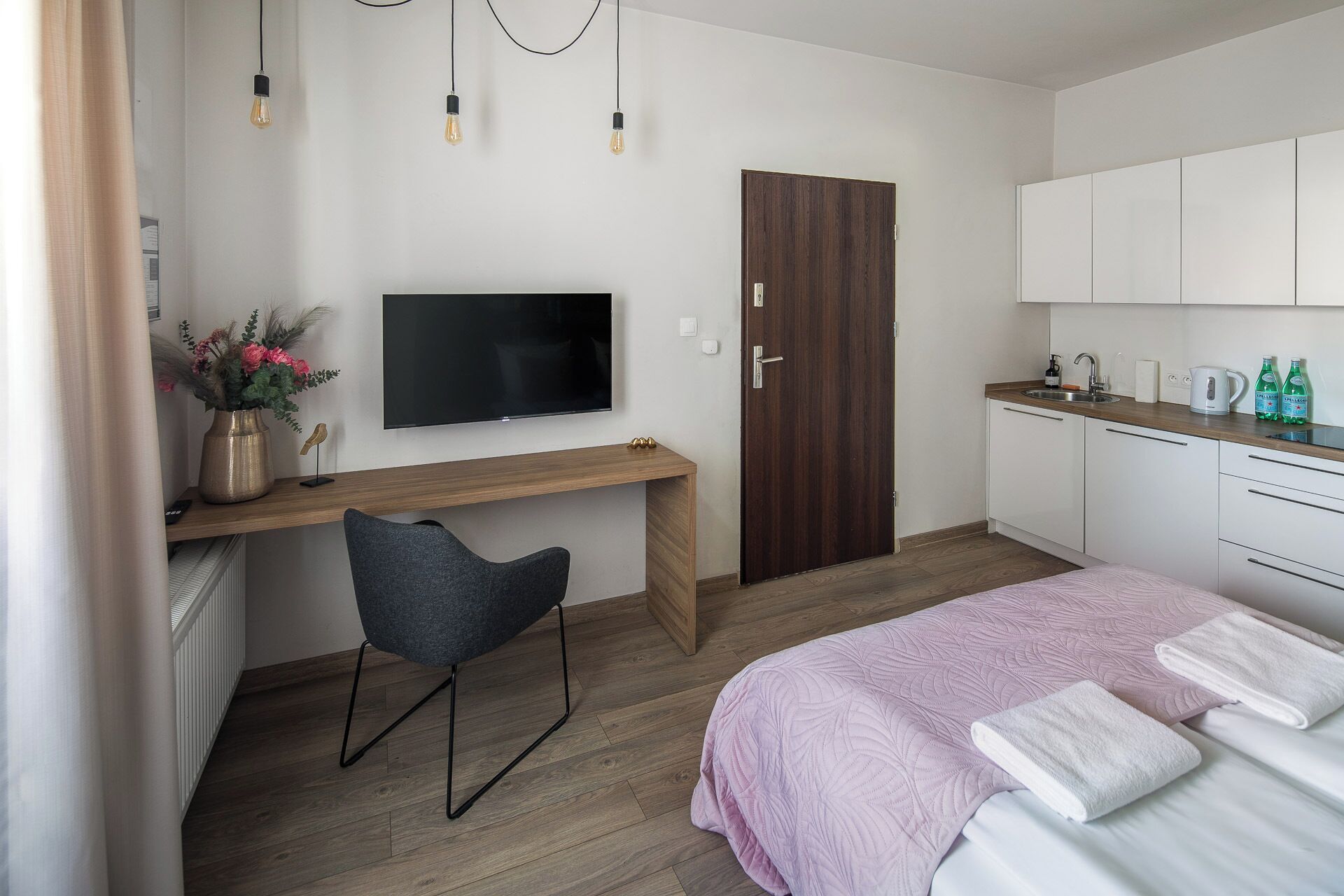 A modern hotel room with a wooden desk, a flat-screen TV, a grey chair, a bed with a pink quilt, and a kitchenette with white cabinets.