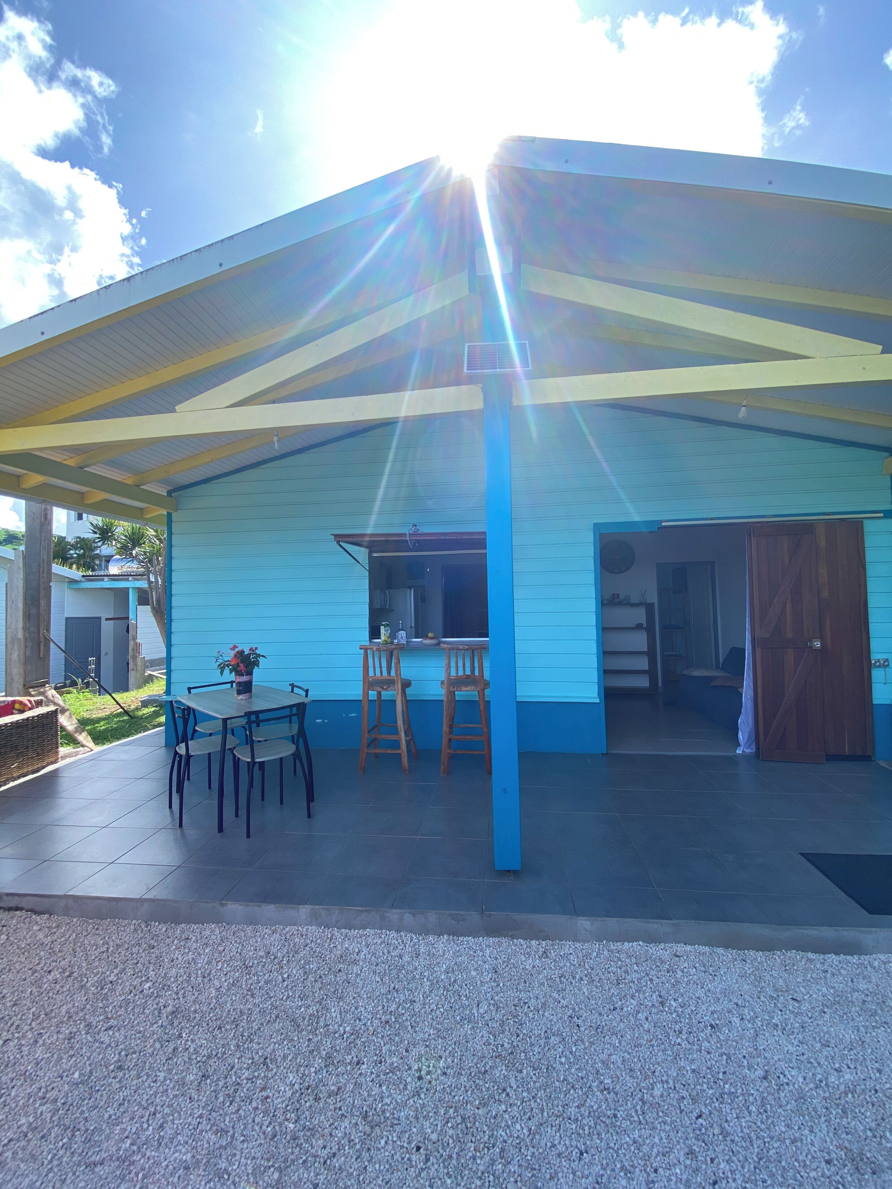Un área de patio techada con una mesa y sillas, un edificio azul al fondo y un cielo despejado.