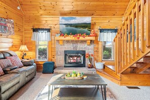 Interior de cabana de toras com lareira, um sofá, mesa de centro e pintura com paisagem.