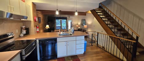 Cocina con isla central, lavavajillas, estufa y pileta. Se ve una escalera de madera que sube a otro nivel.