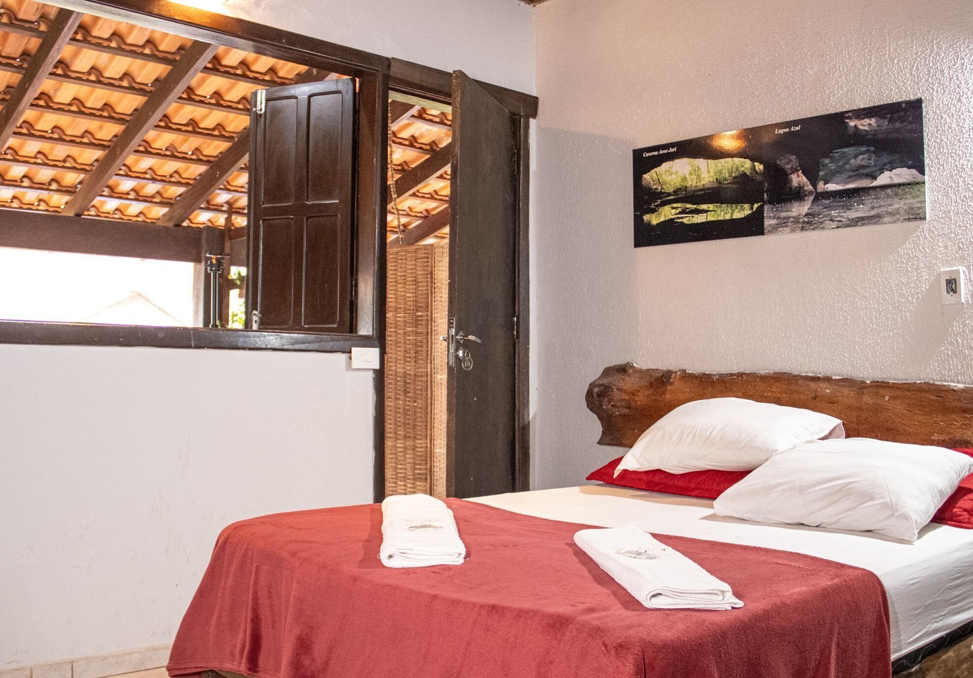 Habitación de hotel con cama, colcha roja, almohadas blancas, cabecera de madera, un cuadro en la pared y una ventana con techo de madera.